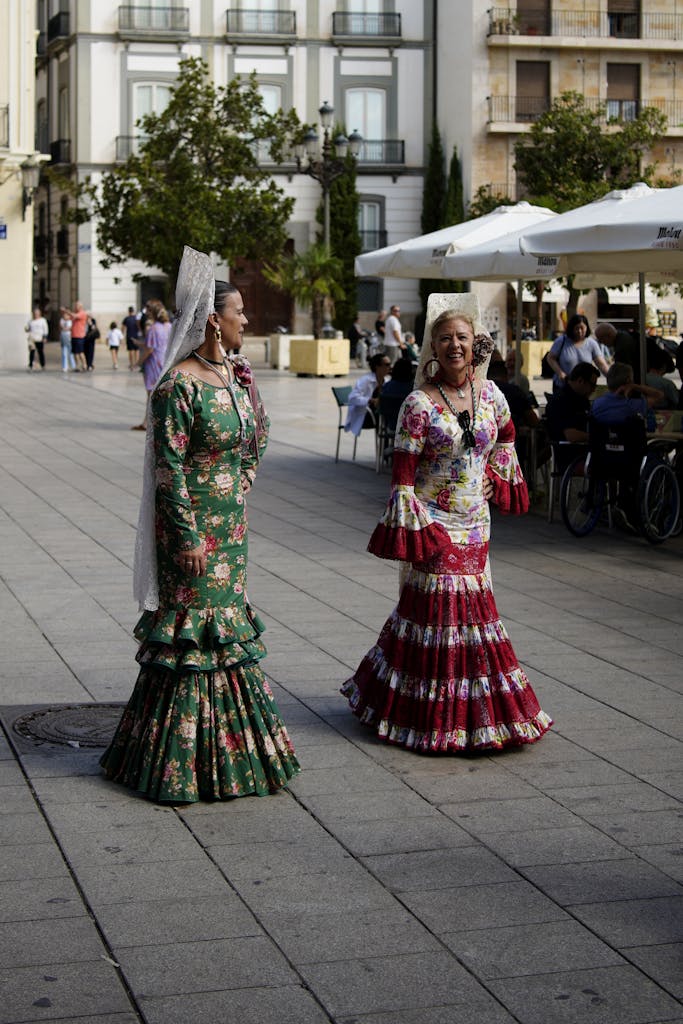 Two women wearing brightly colored flamenco dresses walk down a city plaza.